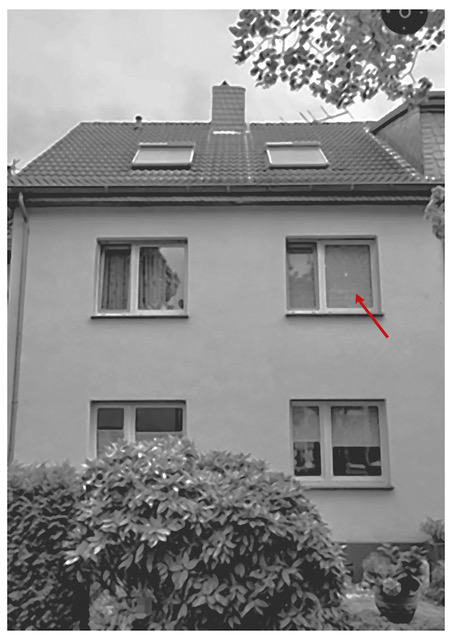 Das Foto (scharz/weiß) zeigt eine zweigeschossige Zweifamilienhaus-Hälfte mit Fenstern in der Dachschräge. Auf das rechte Fenster im zweiten Stock deutet ein roter Pfeil von rechts unten nach links oben.